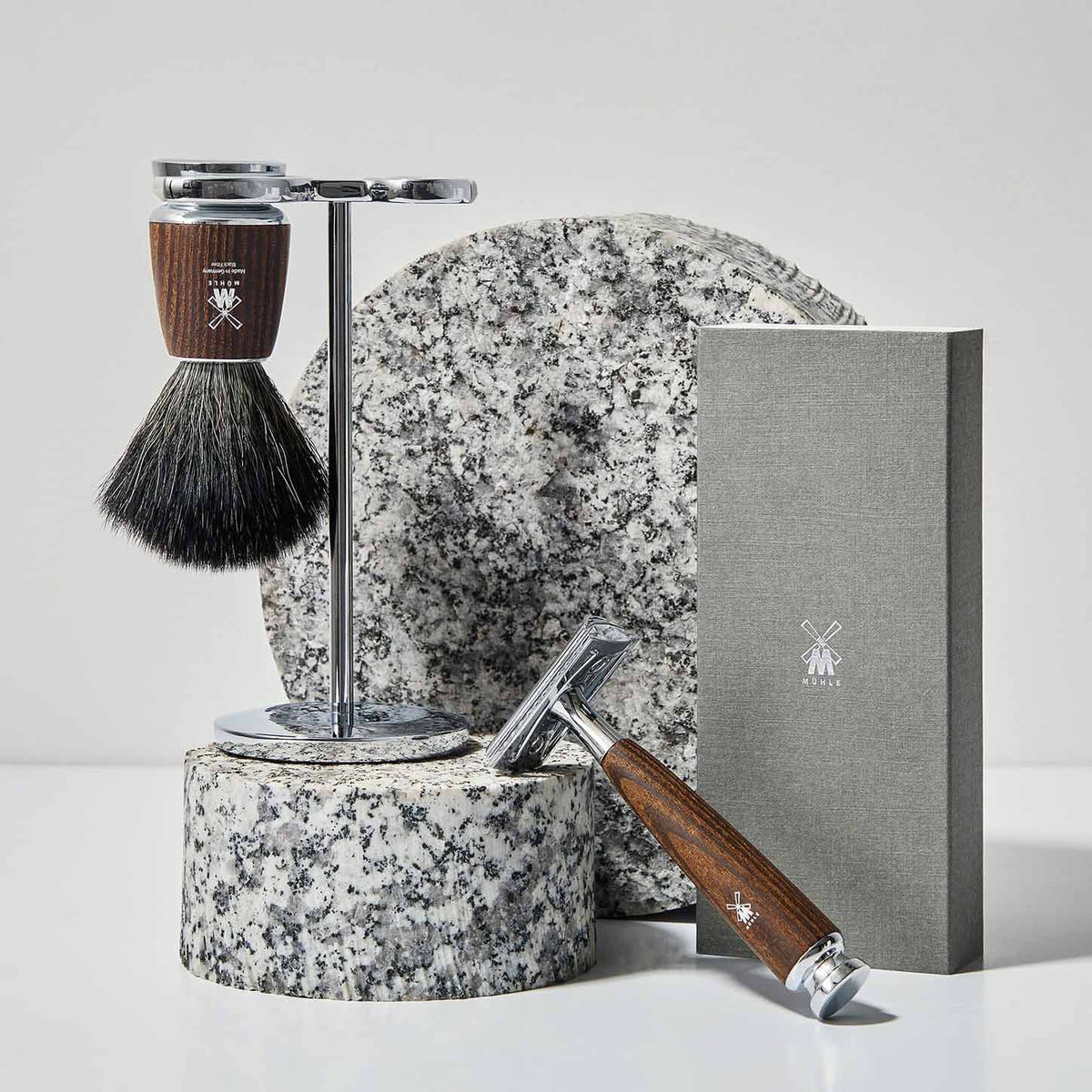 A MUHLE Rytmo Series shaving set, including a shaving brush on a stand, a safety razor with a wooden handle, a round speckled stone pad, and a gray rectangular product packaging box.