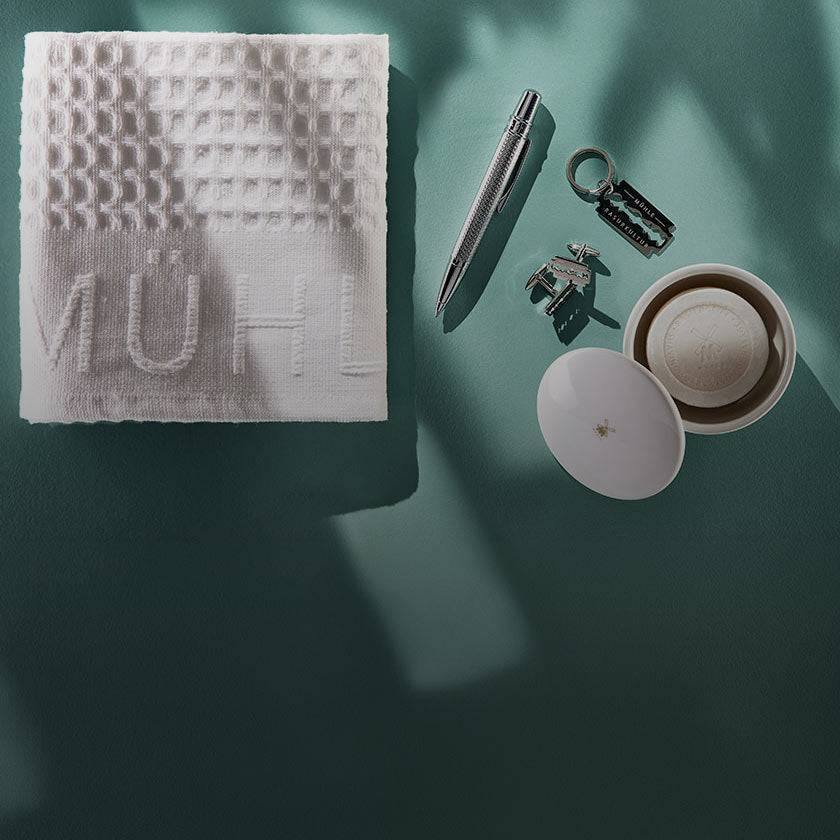 A styled flat-lay of a white textured cloth shaving towel with the brand name 'MÜHLE' embossed on it, arranged alongside a silver pen, razor keychain, and porcelain shaving soap bowl on a dark teal-green background with dramatic shadows.