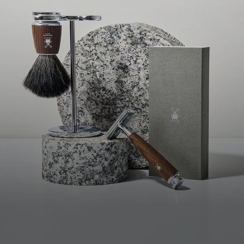 A MUHLE RYTMO Series shaving set arranged on a gray studio surface: a shaving brush on a metal stand, a metal razor with a wooden handle, a gray rectangular box, and a large speckled stone slab behind them.