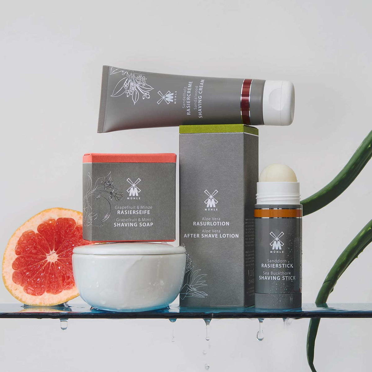 A set of MUHLE shaving products arranged on a shelf with decorative grapefruit and green leaves. The products include a shaving cream tube, a shaving soap, a shaving lotion, a shaving stick, and a small shaving bowl.
